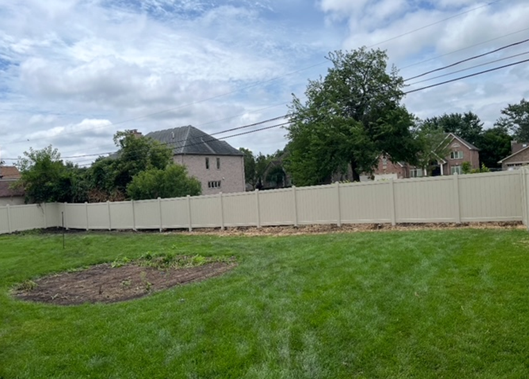 A beige vinyl fence encloses a green backyard, with houses and a cloudy sky in the background. There's a dirt patch in the yard.