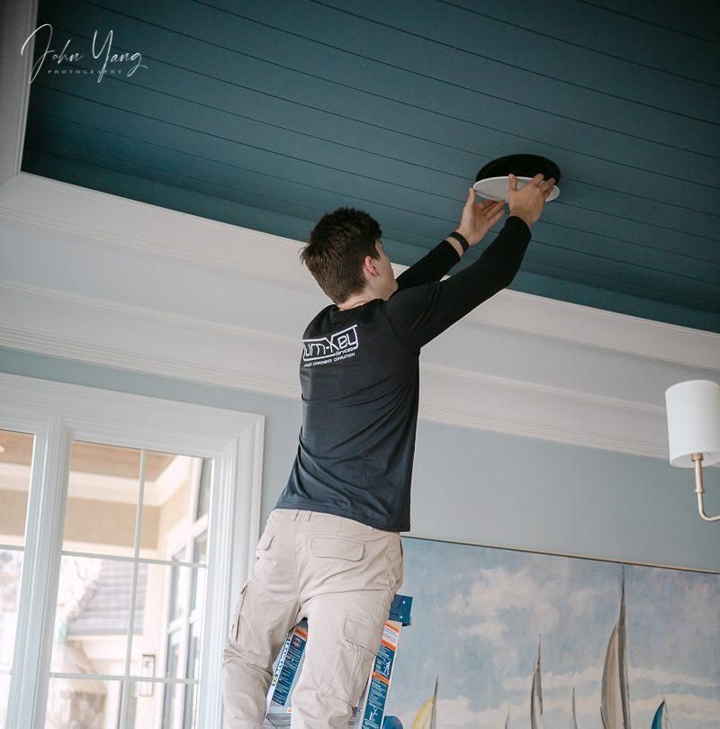 A person installing a ceiling fixture, standing on a ladder in a room with a blue ceiling.