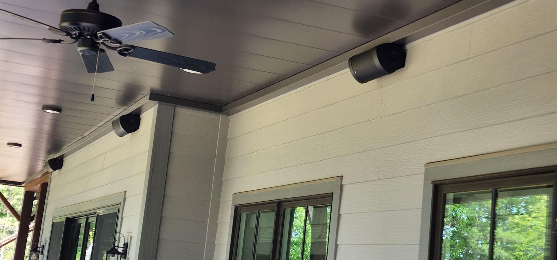 Outdoor patio with speakers and ceiling fan. Grey ceiling, white siding, and windows.