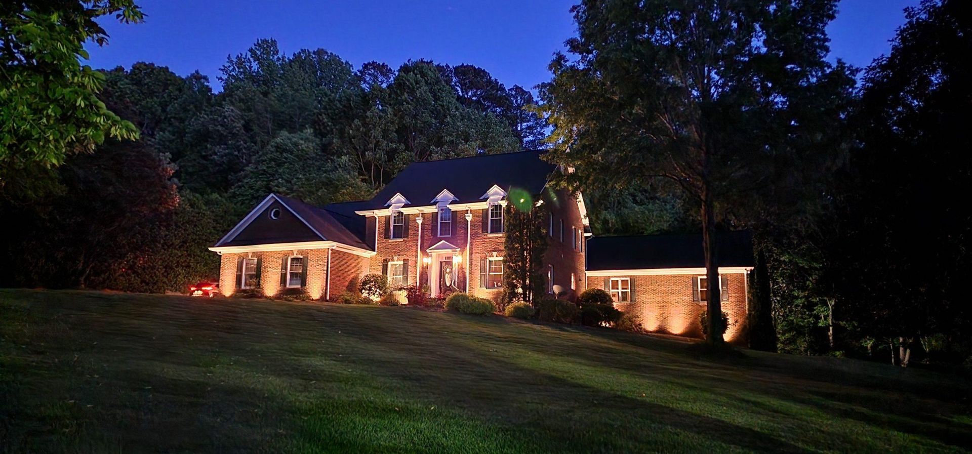 A brick house is illuminated with lights at dusk, with trees in the background and a grassy lawn.