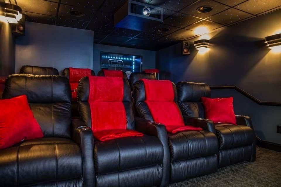 Home theater with black leather recliners, some with red seat cushions and pillows. Dark walls, projector.
