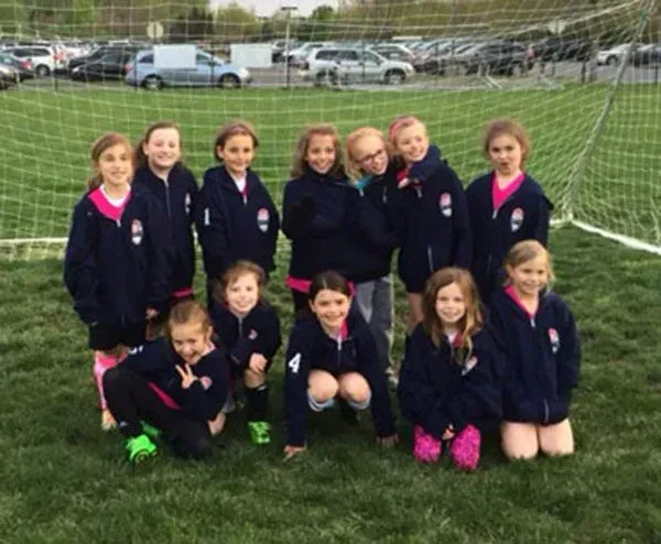Girls soccer team in matching navy jackets on a grassy field, posed in front of a goal.