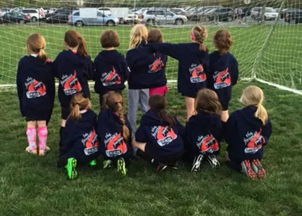 Girls' soccer team in navy blue hoodies with logo on field, backs to the camera.