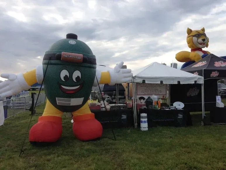 Inflatable grill mascot with arms open, next to a tent and another mascot, outdoors on grass.
