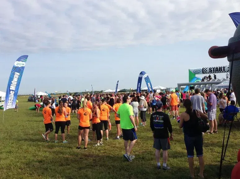 Large crowd at an outdoor race start line under a cloudy sky. People in athletic wear, some wearing orange shirts.