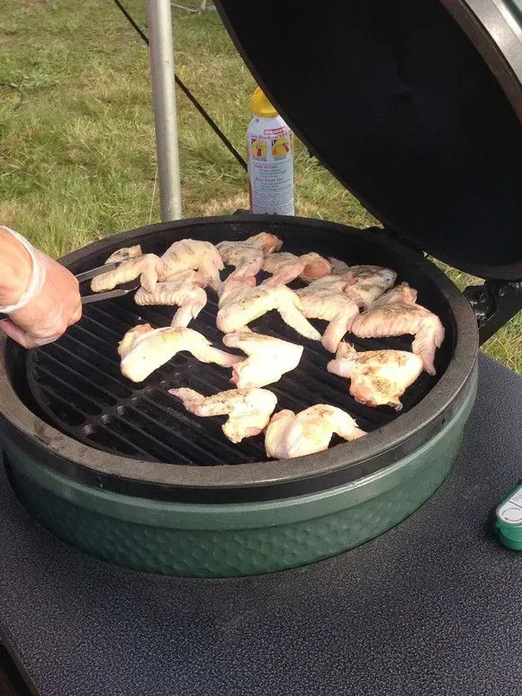 Chicken wings grilling on a green ceramic smoker outside on a sunny day. A person is using tongs.