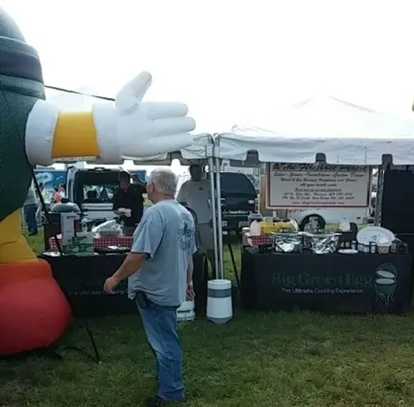 A man walks past a food stand at an outdoor event, featuring a large inflatable mascot and Big Green Egg equipment.