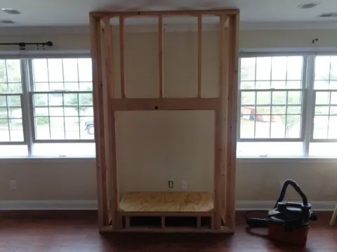 Wooden framework for a fireplace against a beige wall, centered between two windows; vacuum cleaner sits nearby.