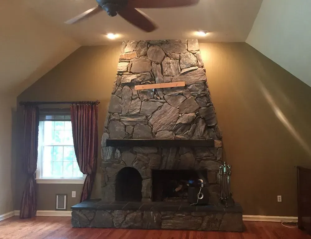 Stone fireplace in a room with maroon curtains, a window, and a ceiling fan.