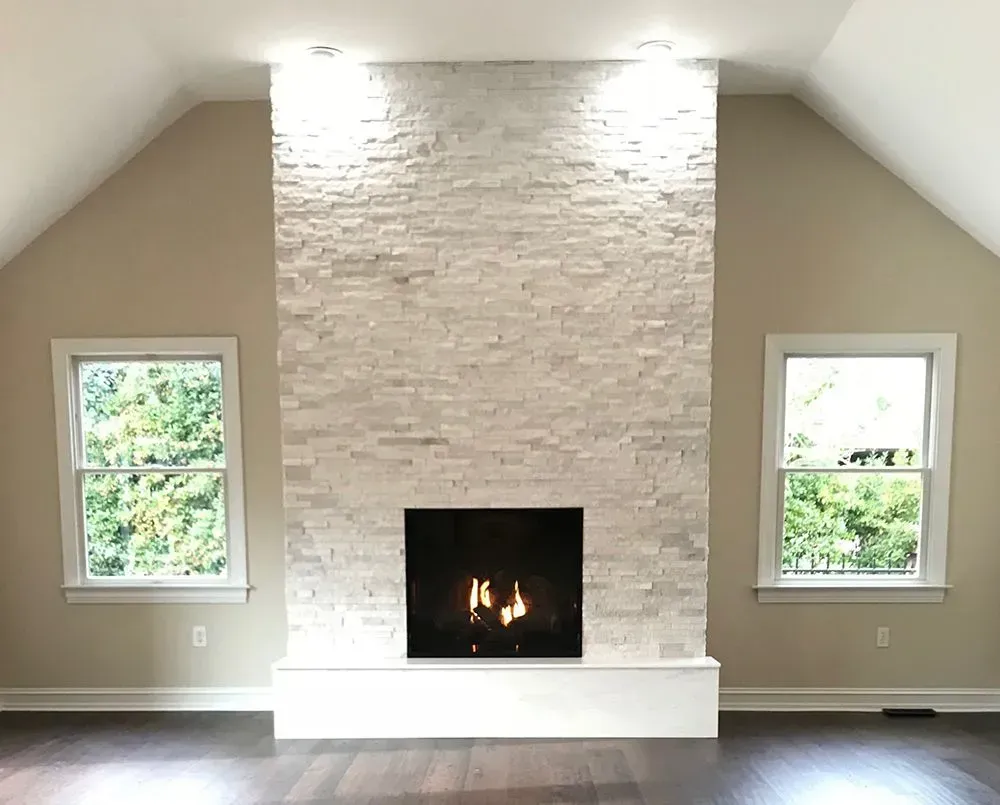 Fireplace with white stone facade, flanked by windows, in a room with vaulted ceiling and dark wood floor.