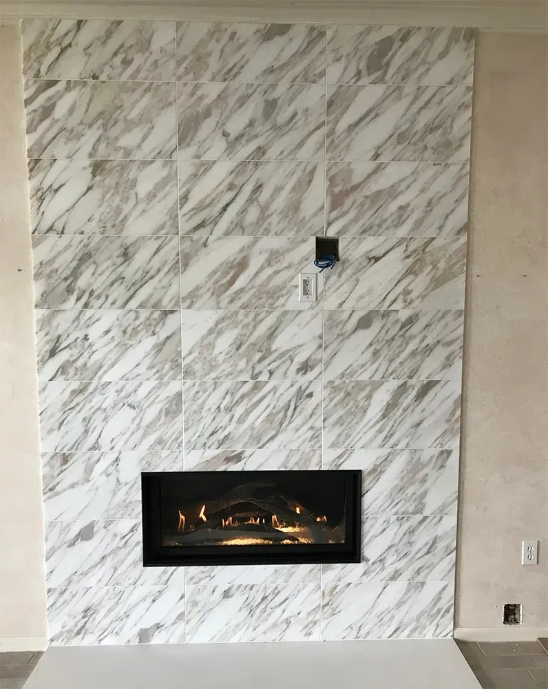 Fireplace with marble-look tile surround. A rectangular firebox glows. The background is light-colored walls.