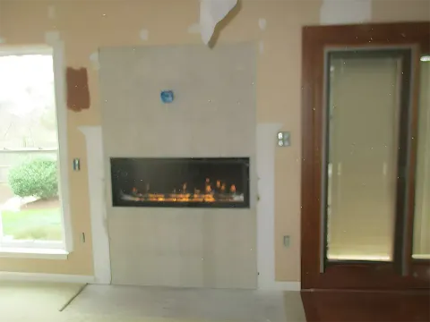 Modern fireplace, recessed in a light-colored wall, with a glowing flame effect. Near a window and wooden door.
