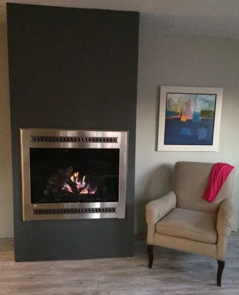 Fireplace with a silver frame on a dark gray wall, next to an armchair with a pink scarf, and a painting.