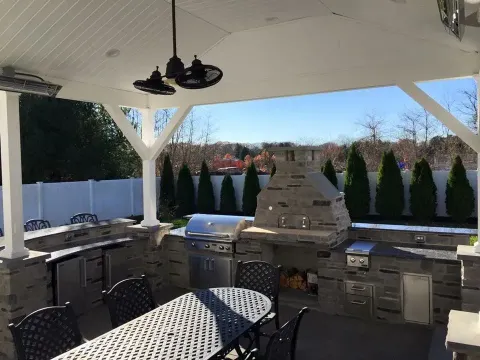 Outdoor kitchen with grill, oven, dining table, and ceiling fan under a white pergola.