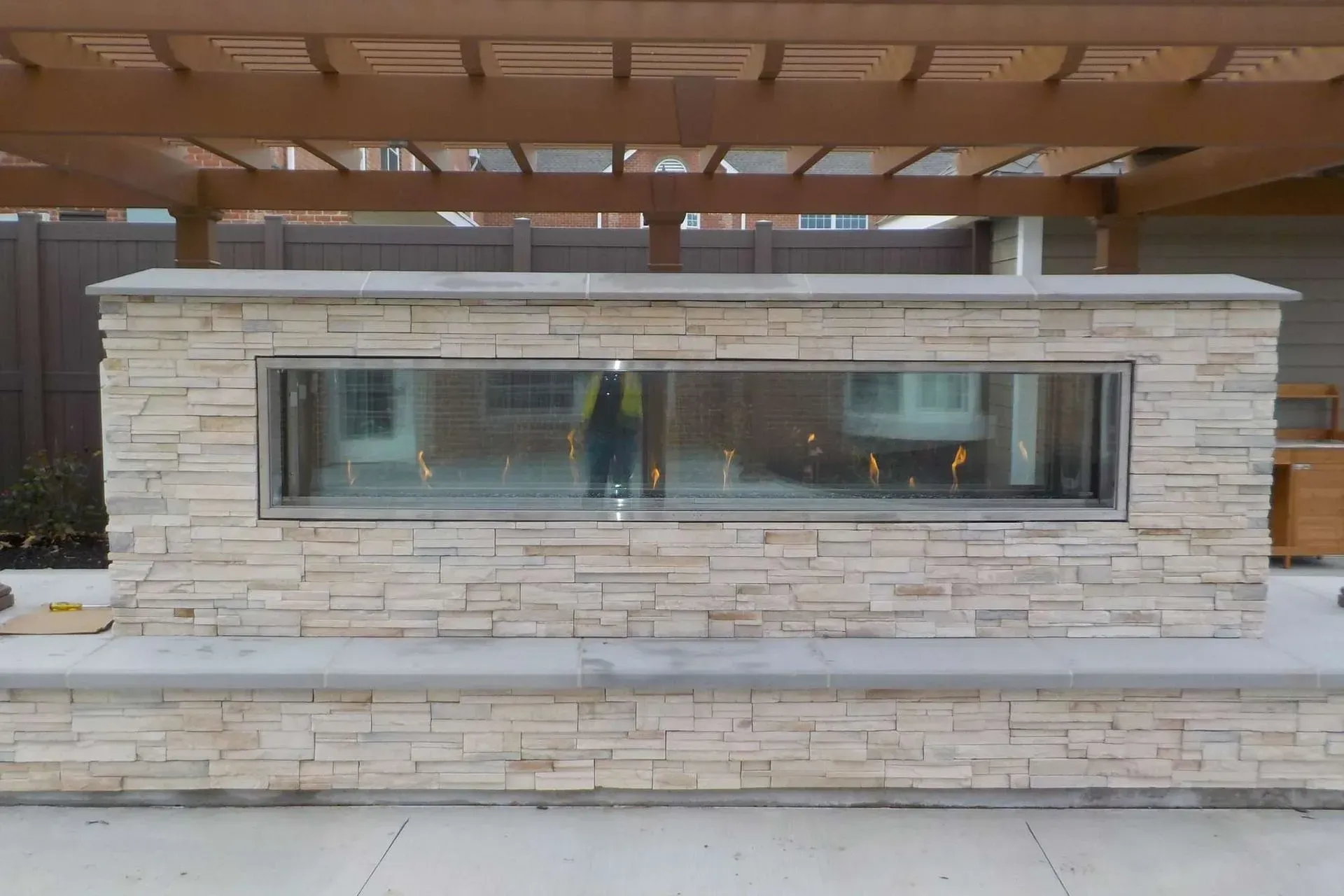 Stone fireplace with a glass front, flames visible, under a wooden pergola.