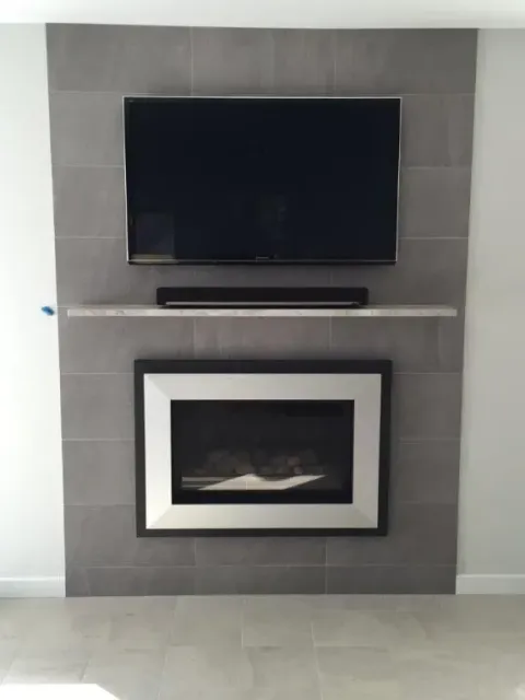 Grey tiled wall with mounted TV, soundbar, and fireplace, accented by a white shelf and silver trim.