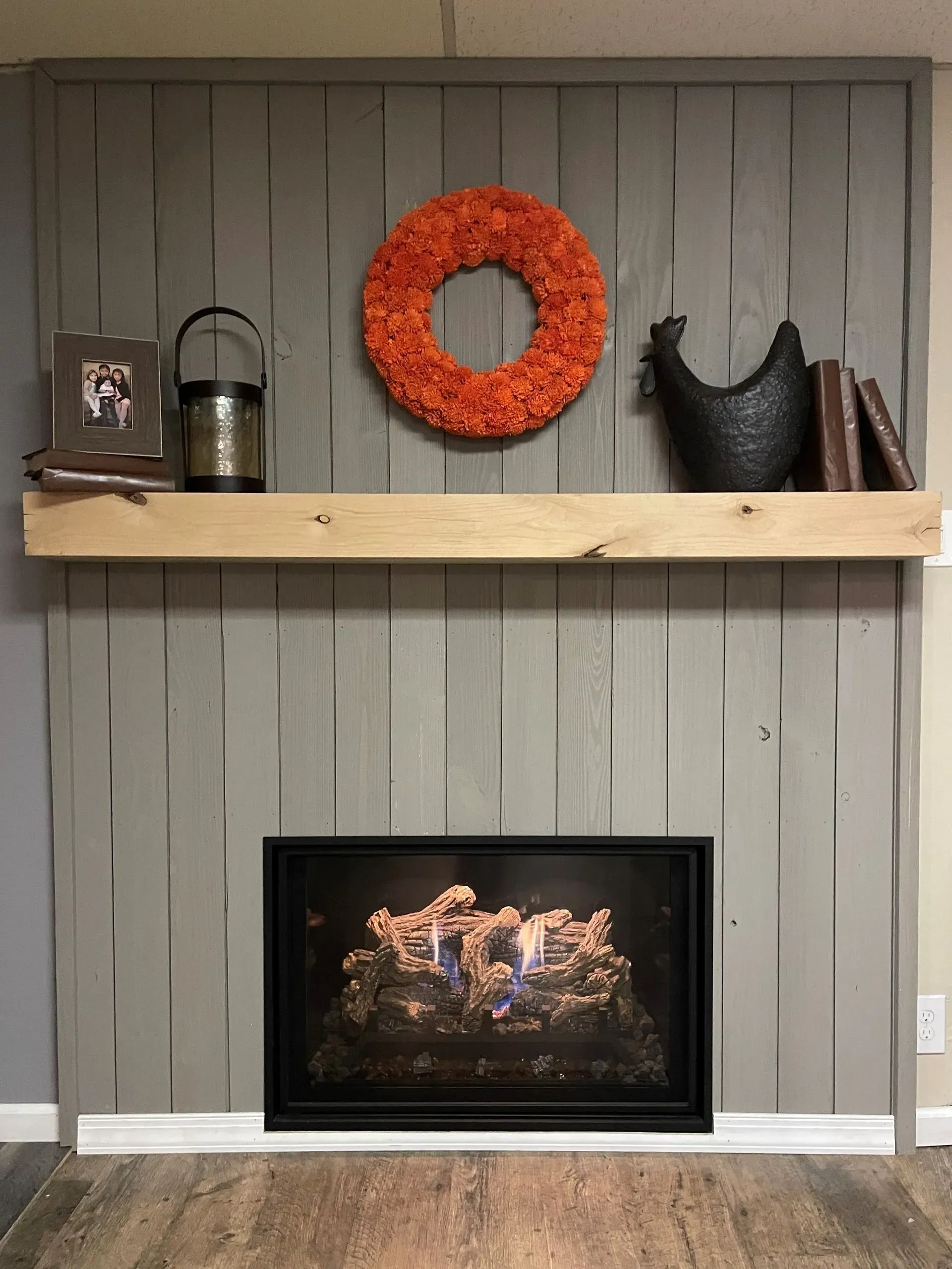 Fireplace with gray wooden paneling, a wooden shelf, orange wreath, and black decor.