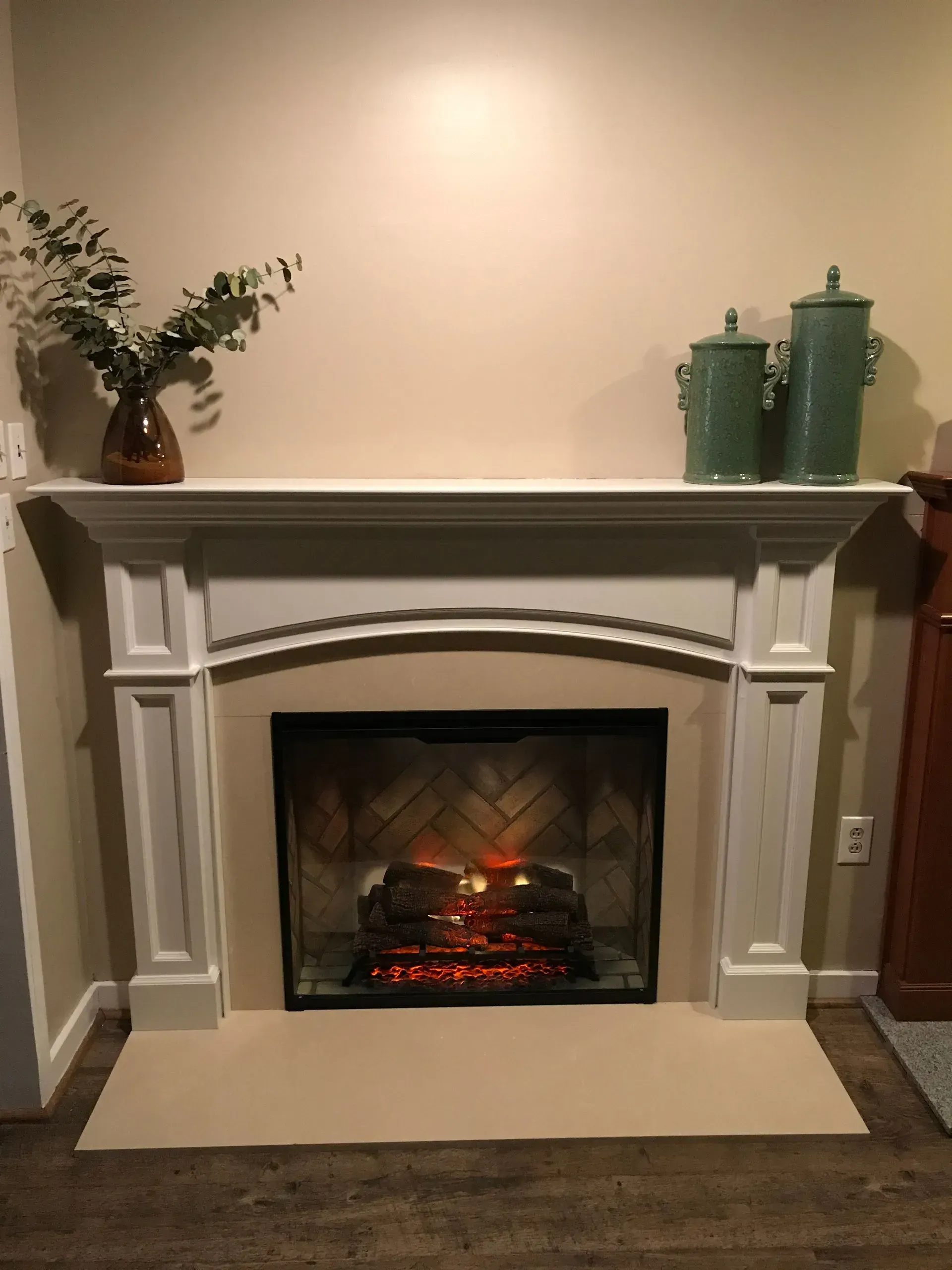 Fireplace with white mantle, beige surround, lit fire, and green decor.