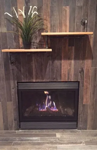 Fireplace with flames, wooden shelves holding plant, against wood-look tiled wall.