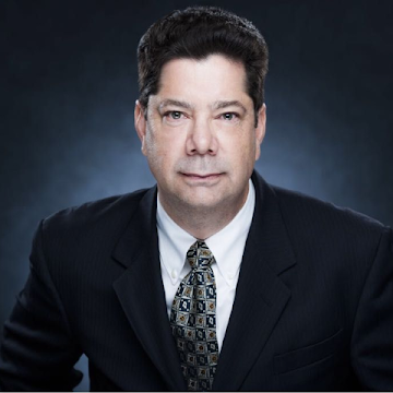 Photo of a person in a dark suit and patterned tie against a dark blue background, looking directly at the camera.