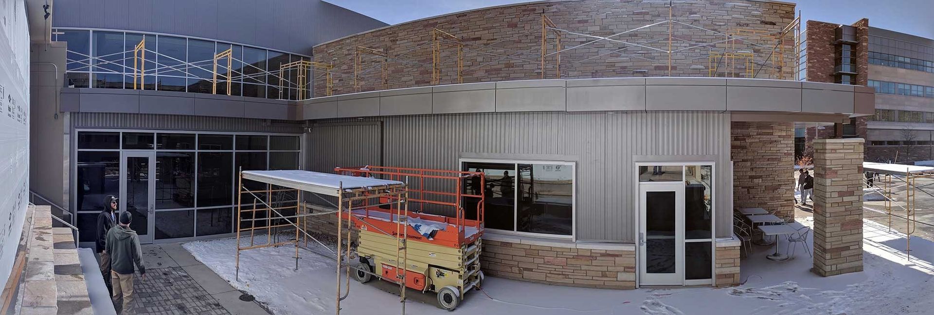 Exterior view of a building under construction, featuring stone and glass details with a lift. Snowy ground is visible.