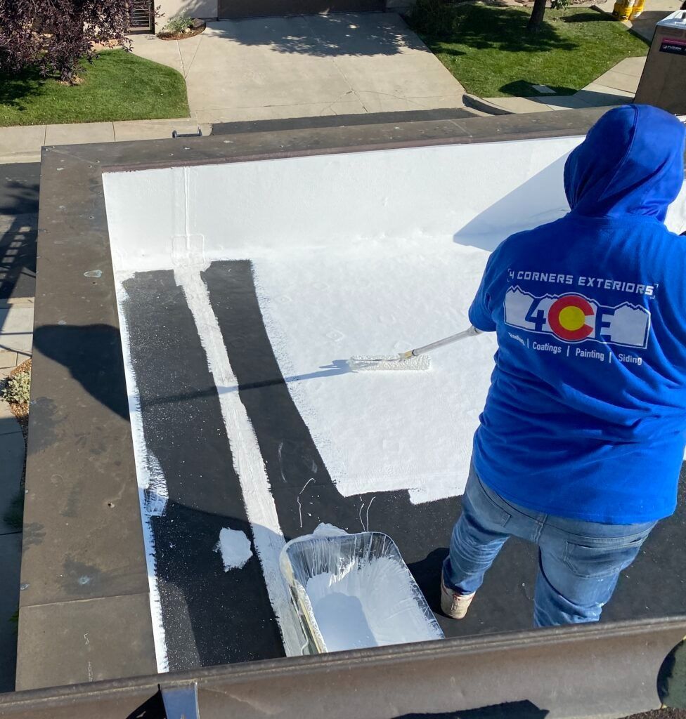 Person applying white coating to a flat roof with a roller. They wear a blue hoodie and jeans, outdoors.