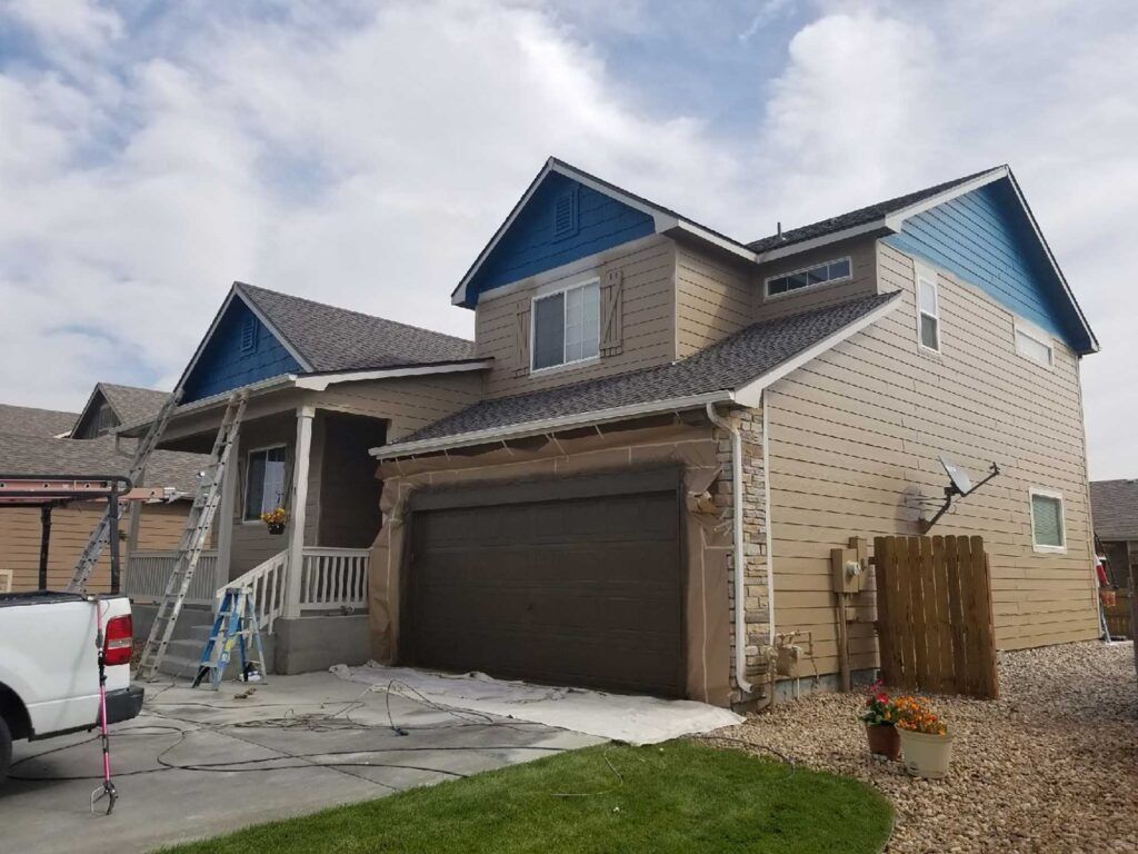 Two-story house with brown garage door, blue roof accents, and tan siding on a cloudy day.