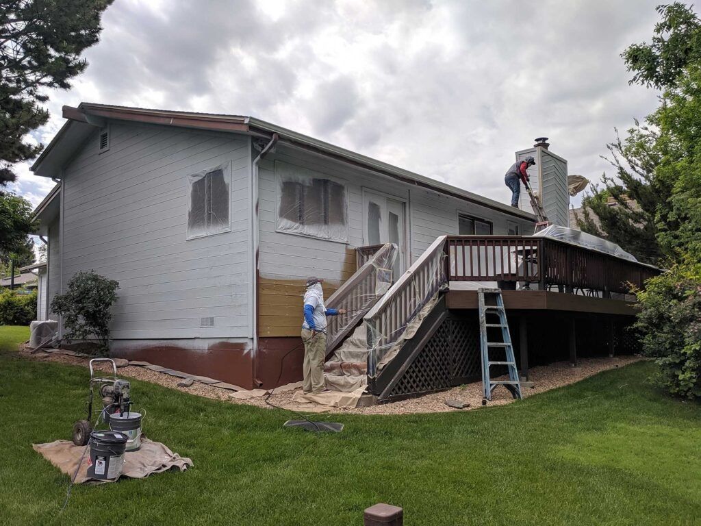 Workers painting a house. One is on the roof, the other on the ground near the stairs. Overcast day.