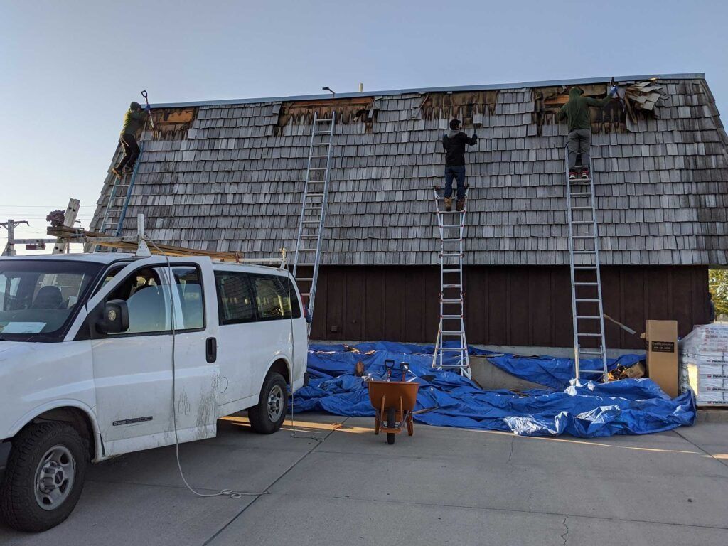 Roofers on ladders replacing shingles on a building with a white van parked nearby.