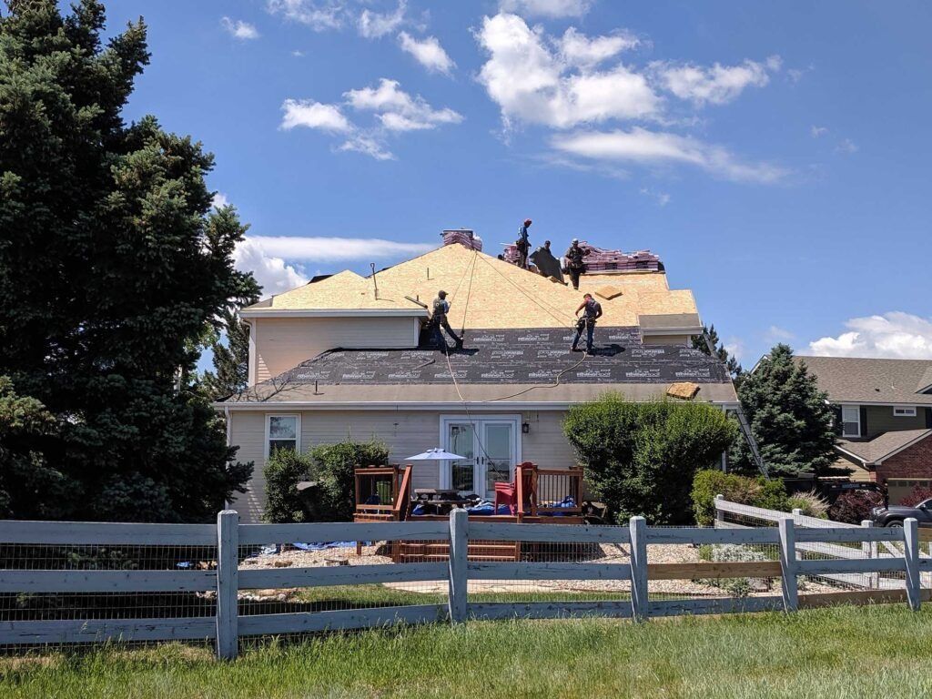 Workers installing shingles on a house roof on a sunny day. White picket fence in foreground.
