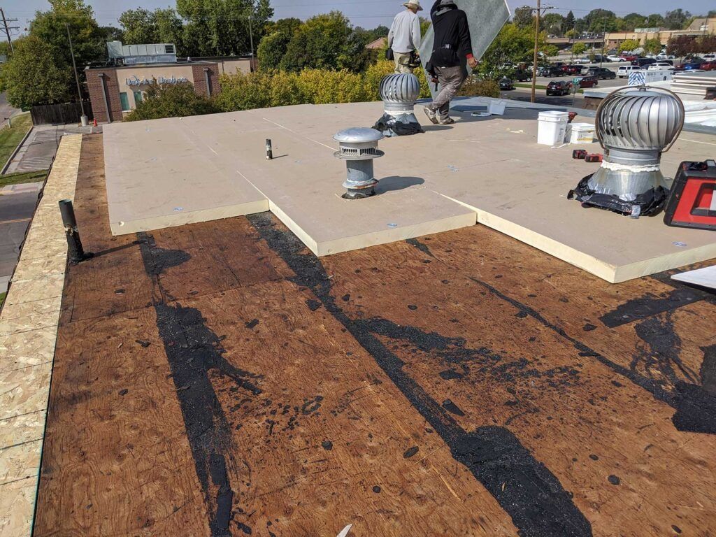 Roofers installing insulation on a flat roof. Brown wood, tan insulation, and silver vents.
