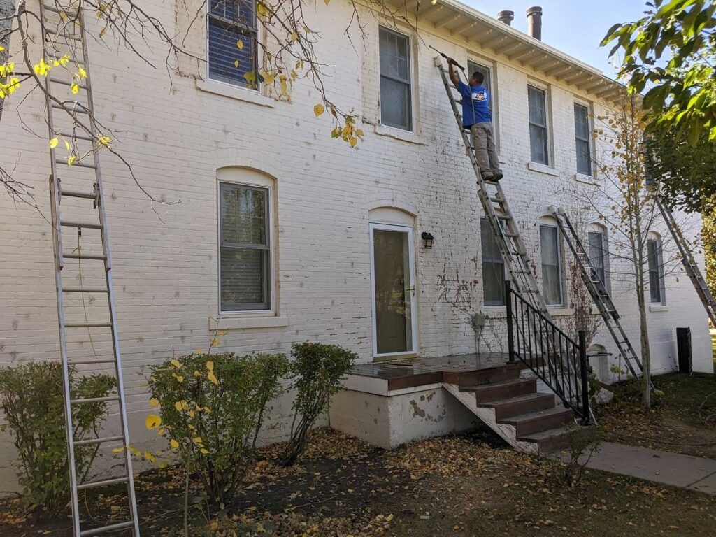Person on ladder painting a two-story white brick building. Multiple ladders set up; a fall day setting.