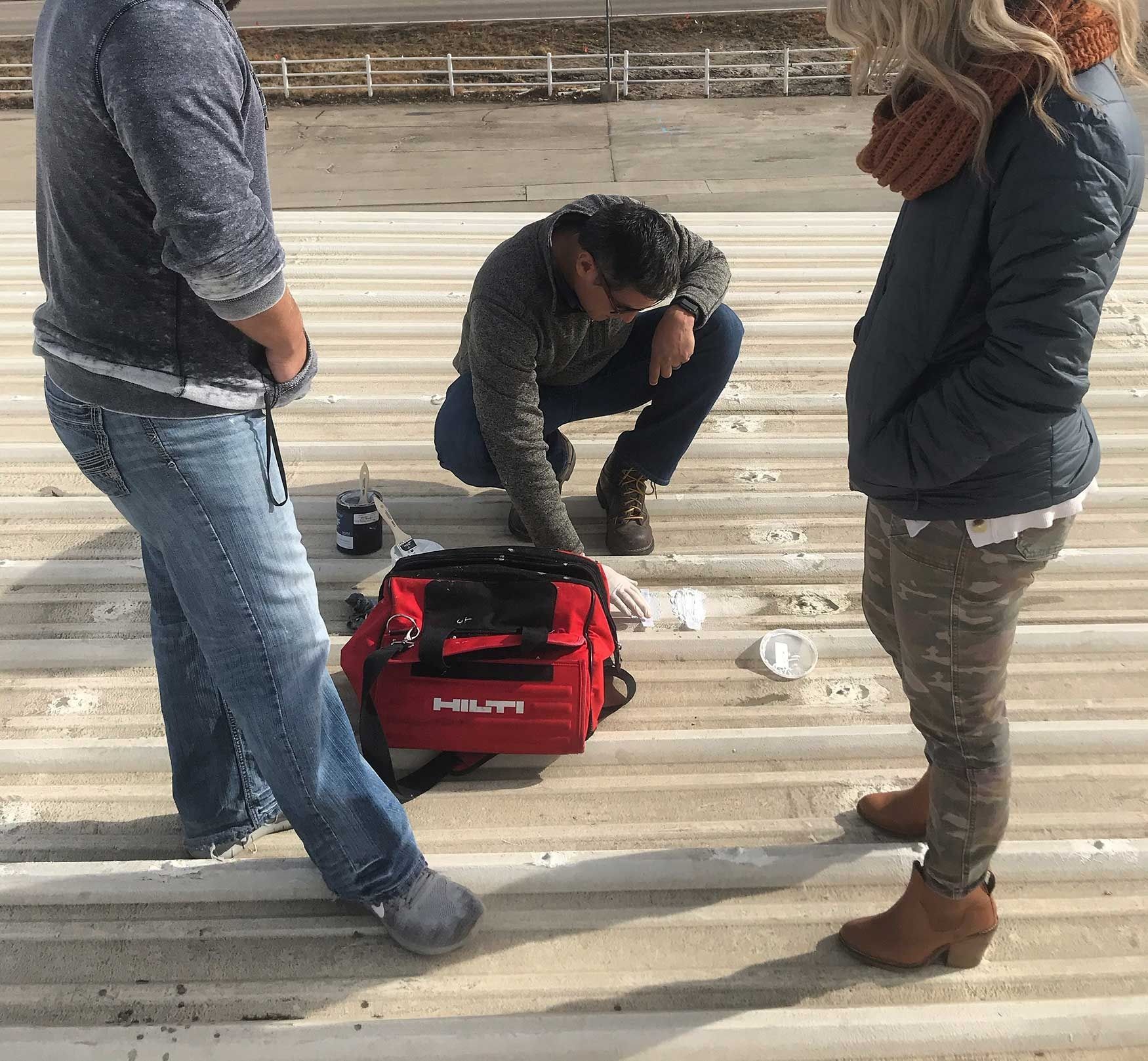 Three people on a corrugated metal roof, one squatting, looking in a red tool bag.