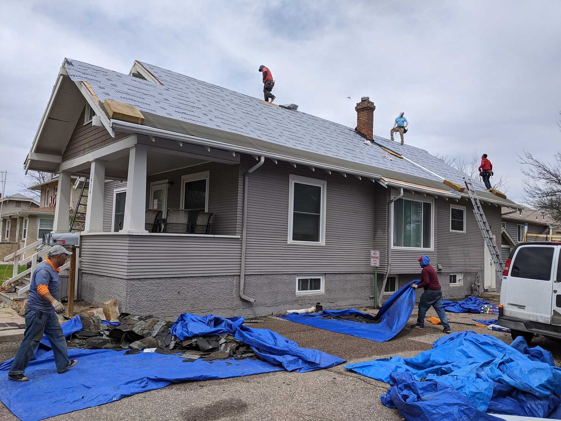 Roofers working on a house with gray siding. Blue tarps cover the ground. Cloudy sky.