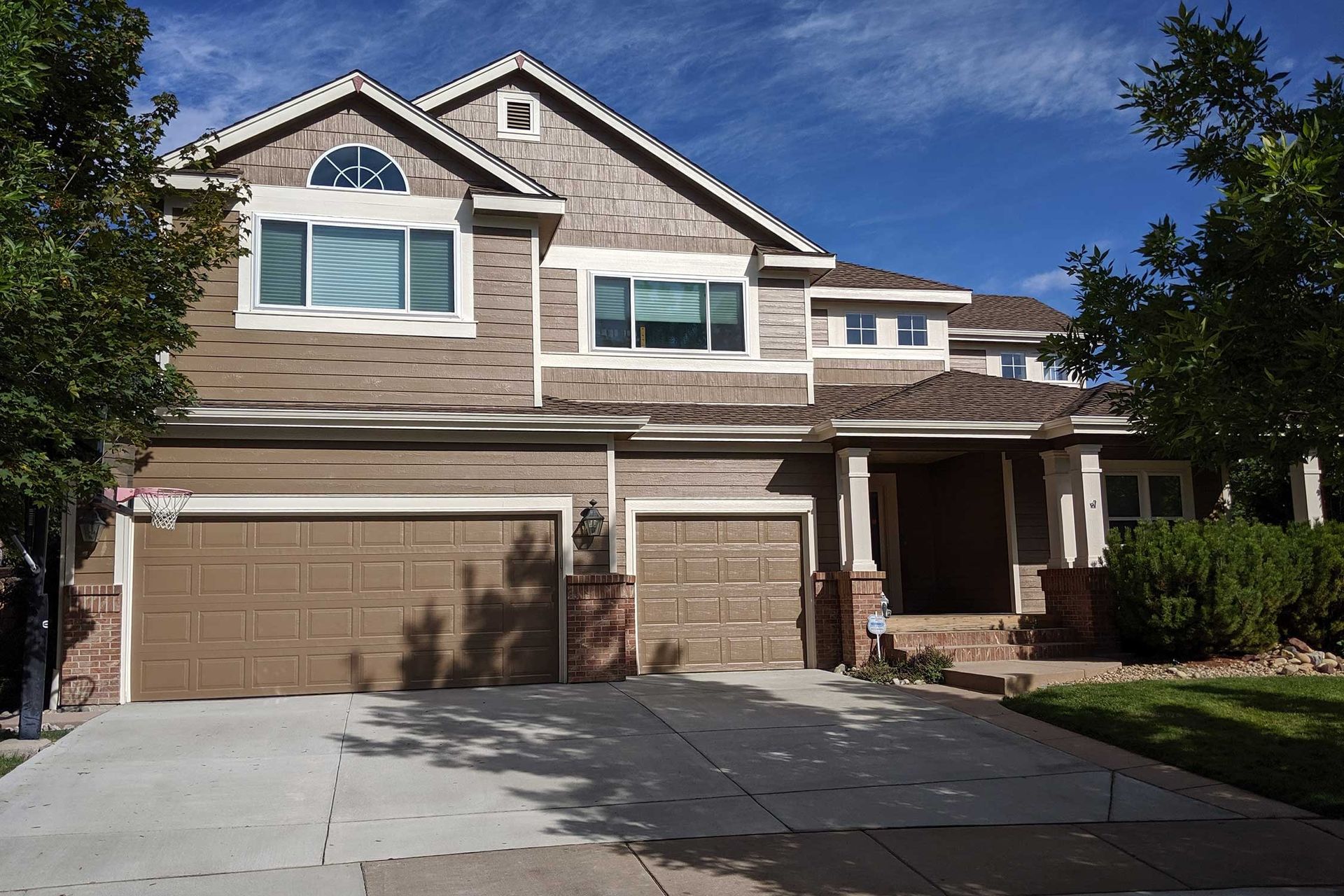 Two-story brown house with a two-car garage, driveway, and front porch. Sunny day, trees present.