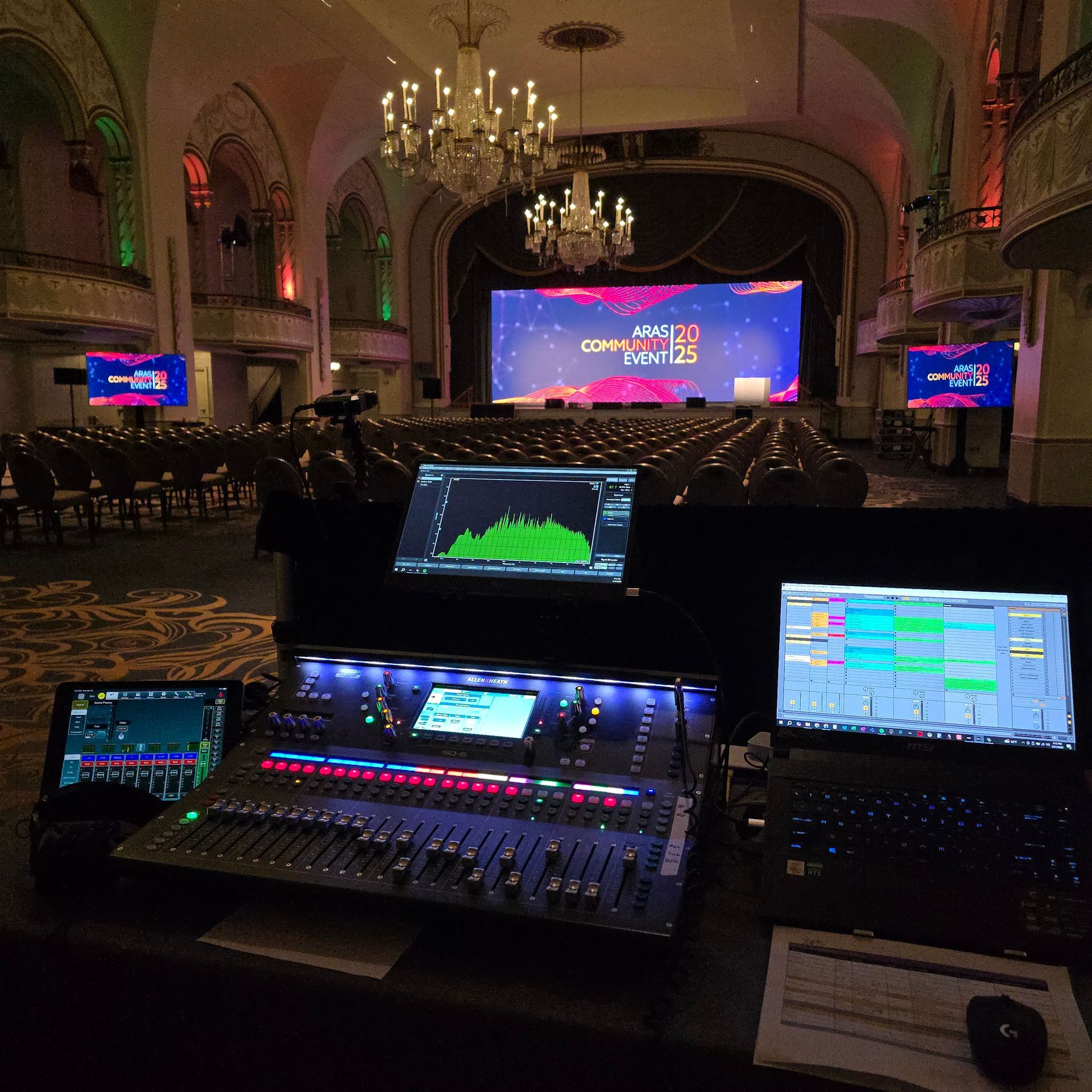 Live event control booth with audio mixing consoles and monitors facing a lit stage in a grand hall