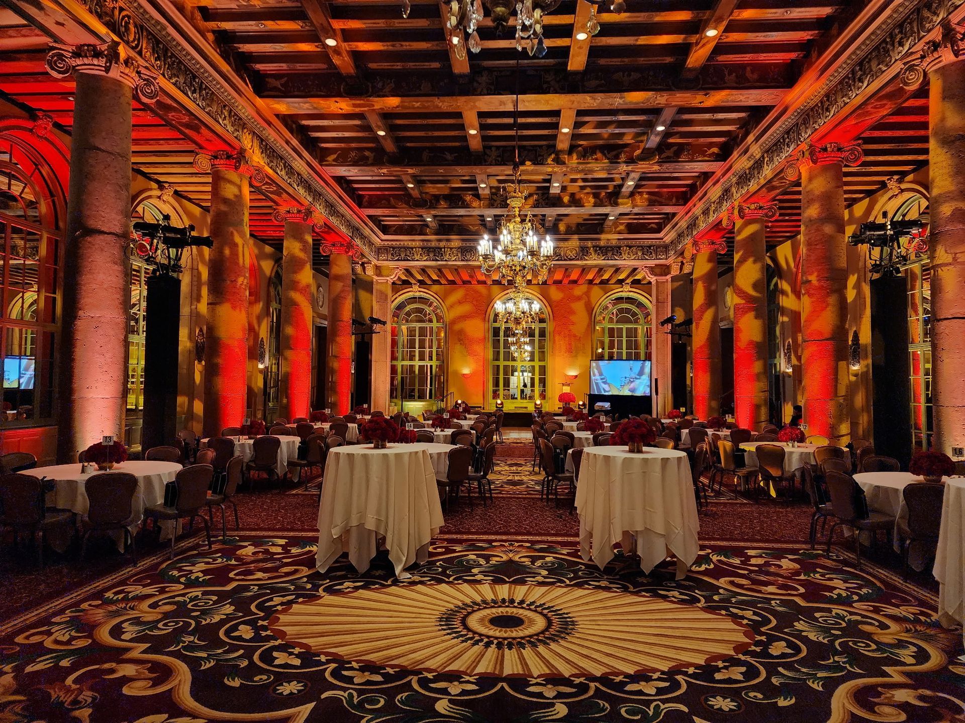 Opulent ballroom with red-lit columns, ornate ceiling, chandeliers, and round tables on patterned carpet