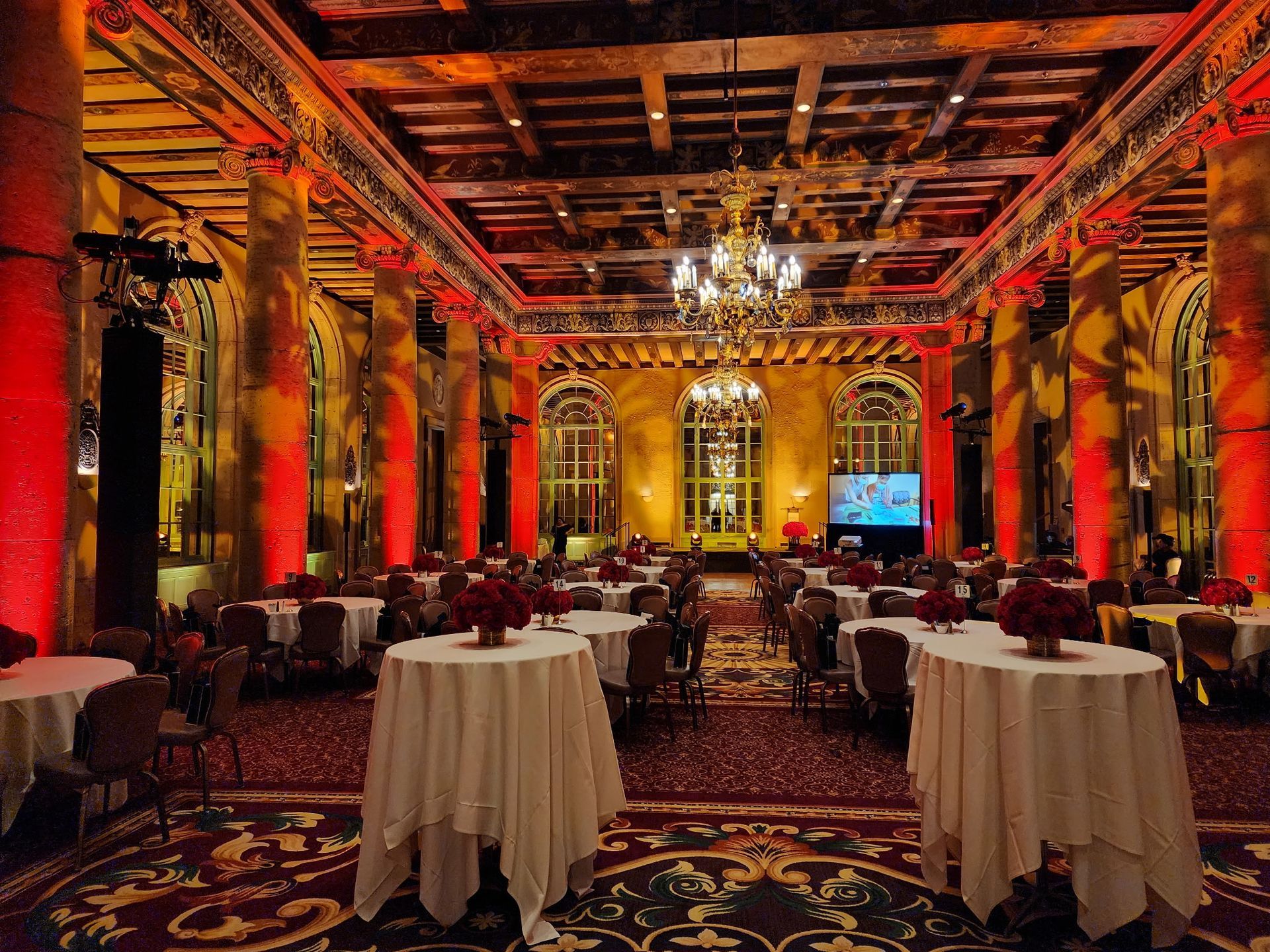 Grand ballroom with red columns, ornate ceiling, and round tables set for an event