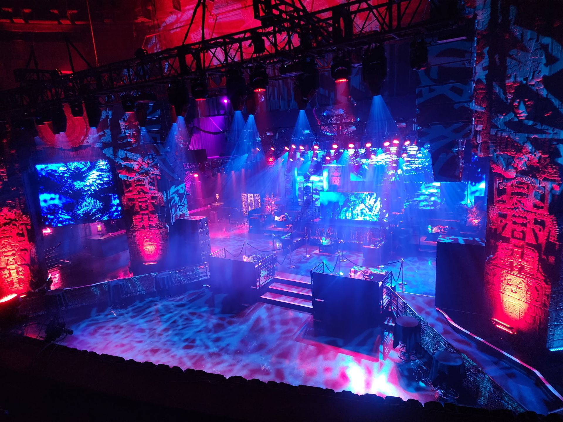 Neon-lit concert stage with blue and pink lights, large LED screens, and a central DJ booth