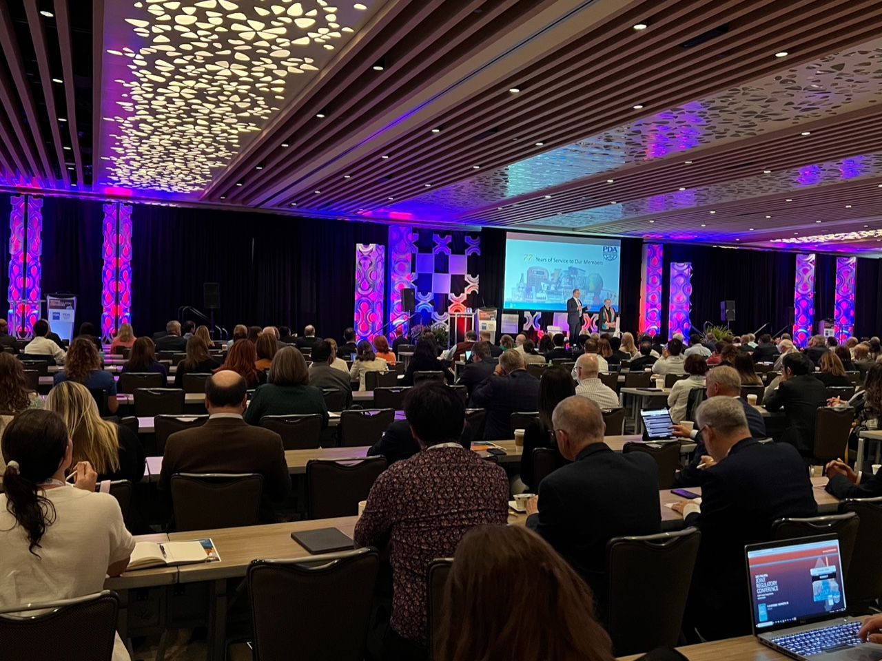 Conference audience seated in a large, purple-lit ballroom facing a presentation screen
