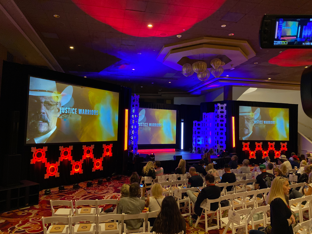 Conference ballroom with seated audience facing three large presentation screens and red-blue stage lighting