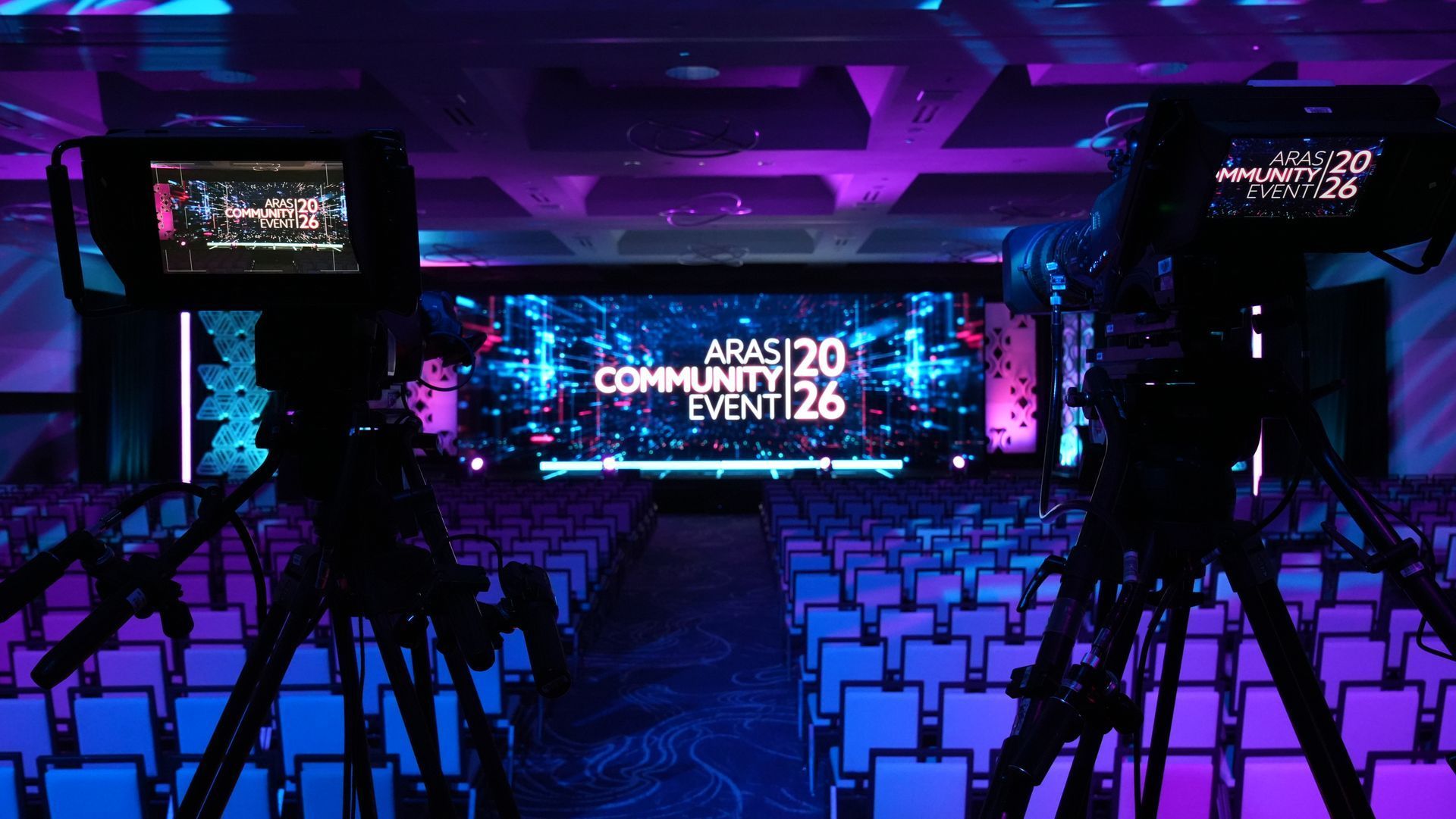 Conference hall with rows of empty white chairs, purple lighting, and a large stage screen reading “AWS Community Event ’26”