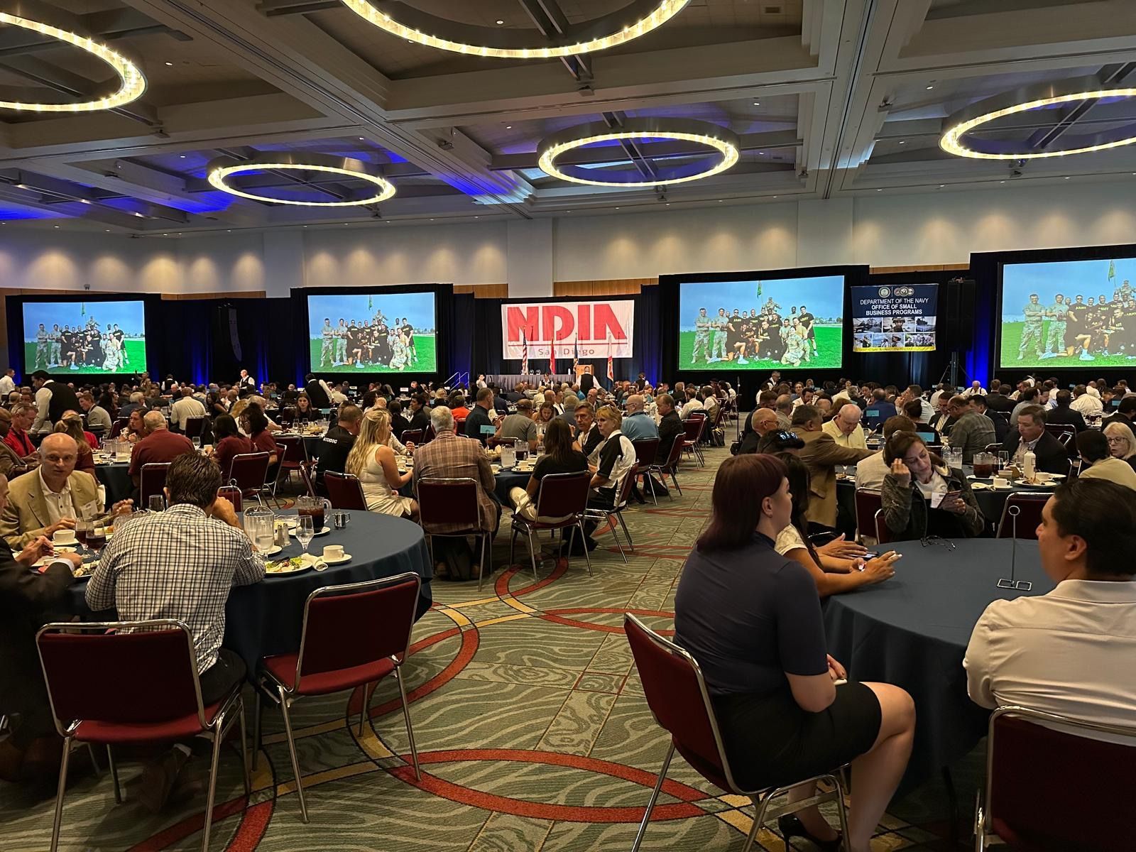 Conference ballroom with seated attendees, stage screens, and overhead ring lights