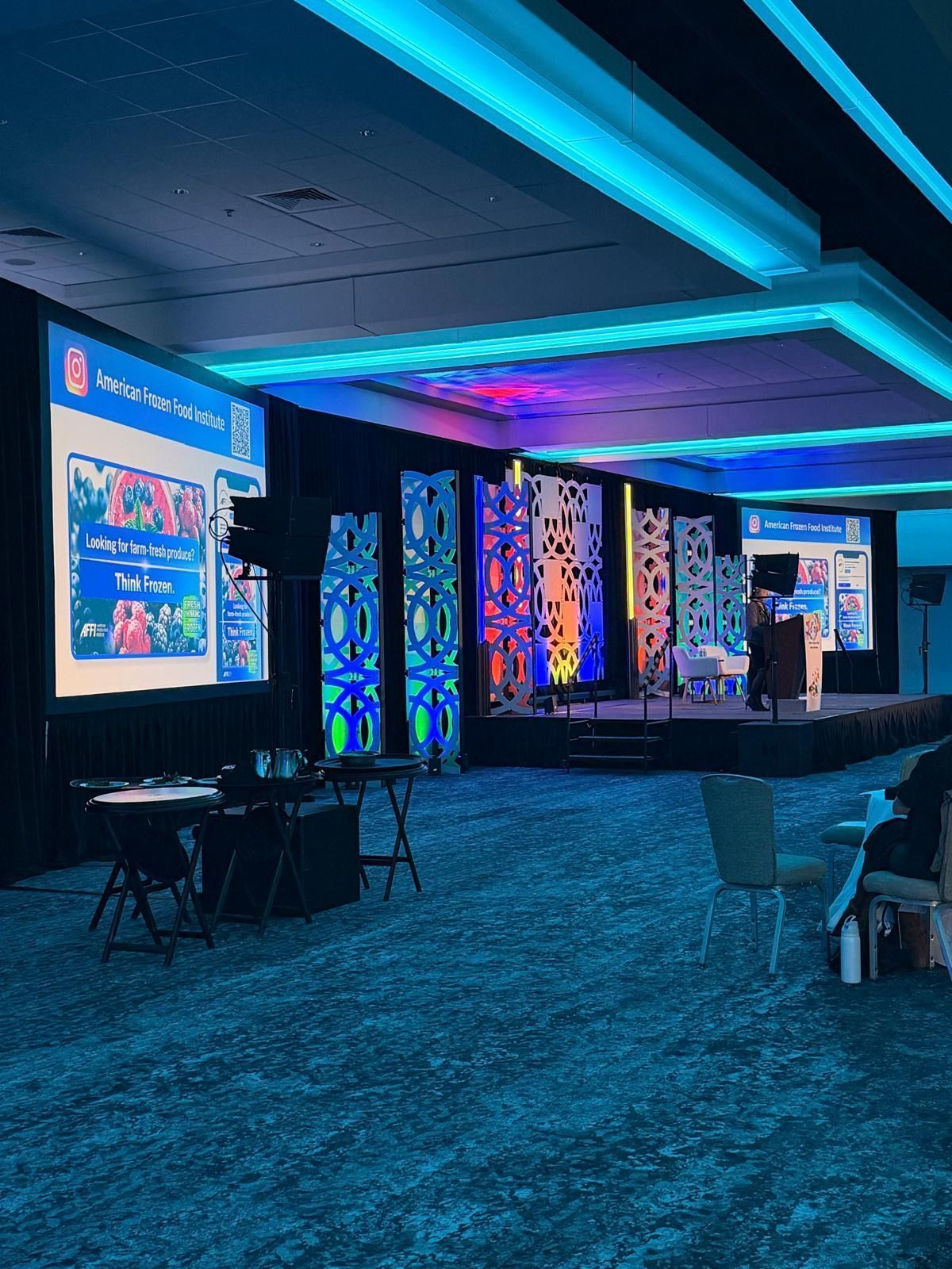 Neon-lit conference hall with stage screens, patterned wall panels, and seating on a carpeted floor