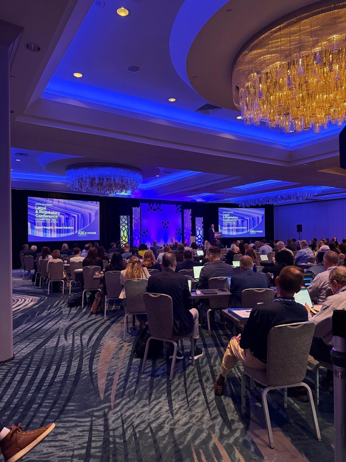 Conference audience seated at round tables in a blue-lit ballroom facing two large presentation screens.