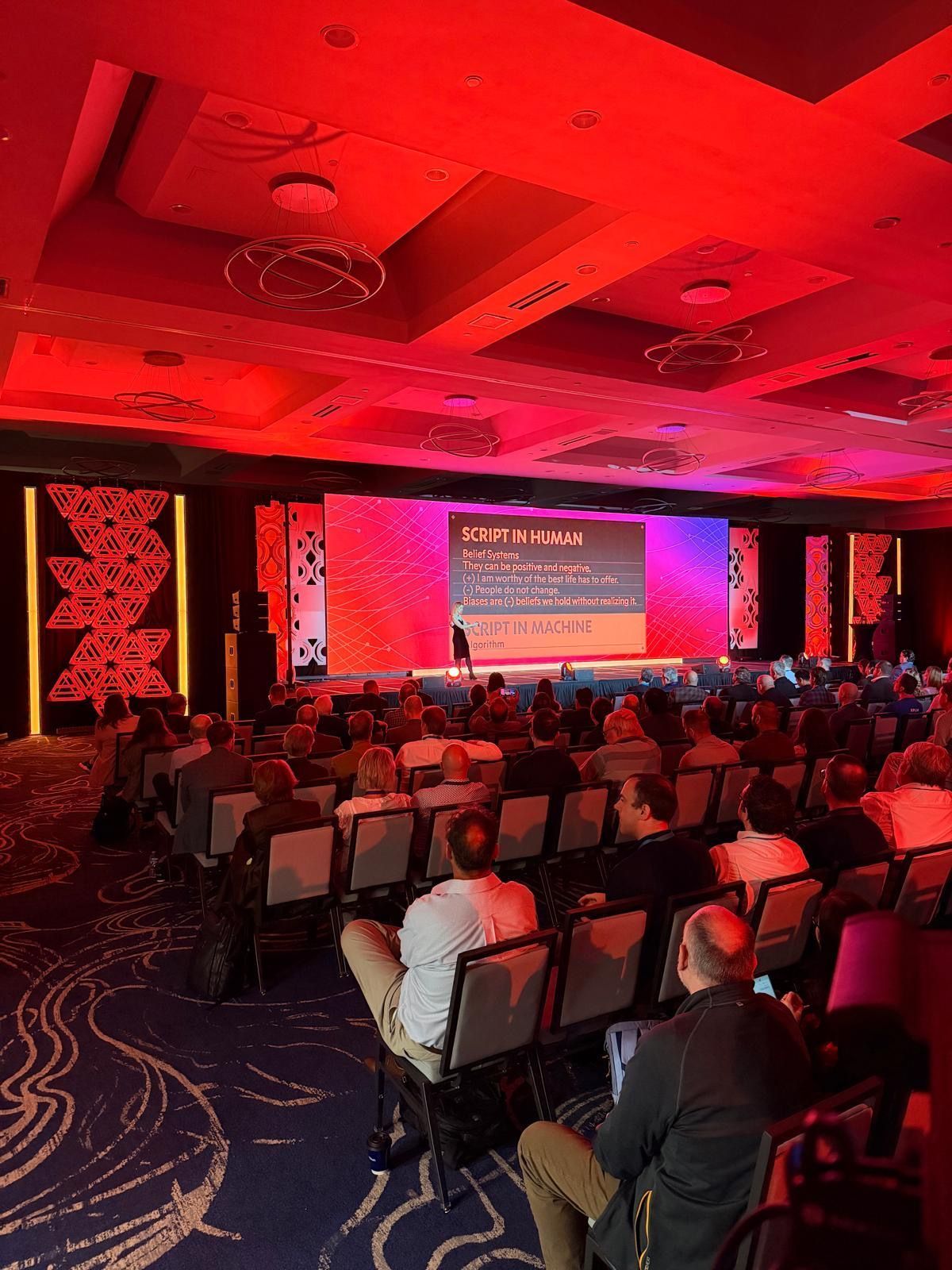Red-lit conference hall with seated audience facing a presentation screen and stage