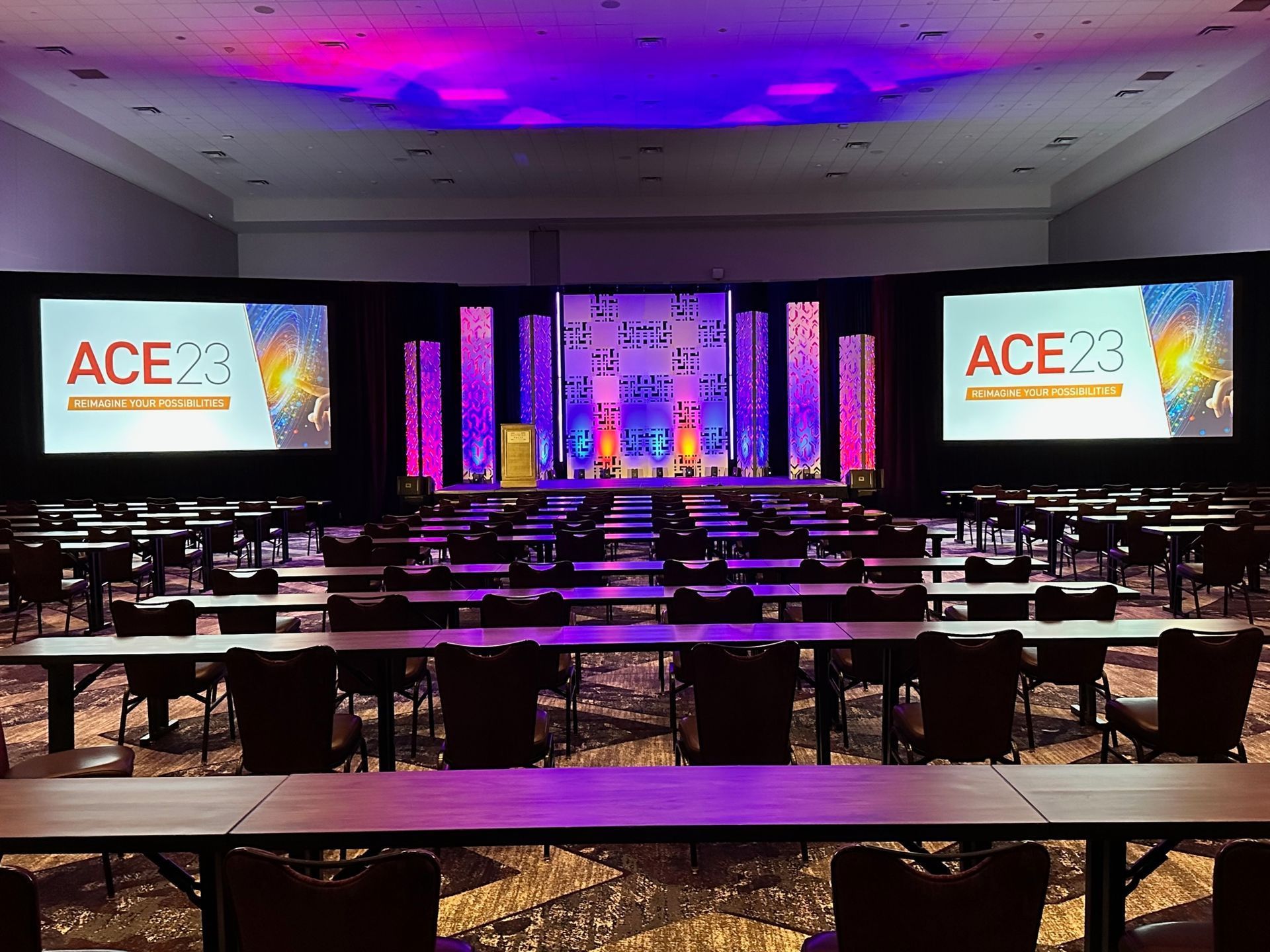 Conference hall with ACE stage and large screens, lit in purple and blue, rows of empty tables in front.