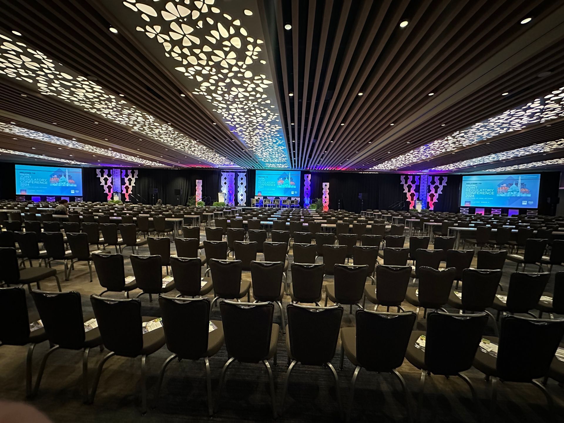 Large conference hall with rows of empty chairs, stage screens, and colorful ceiling lights