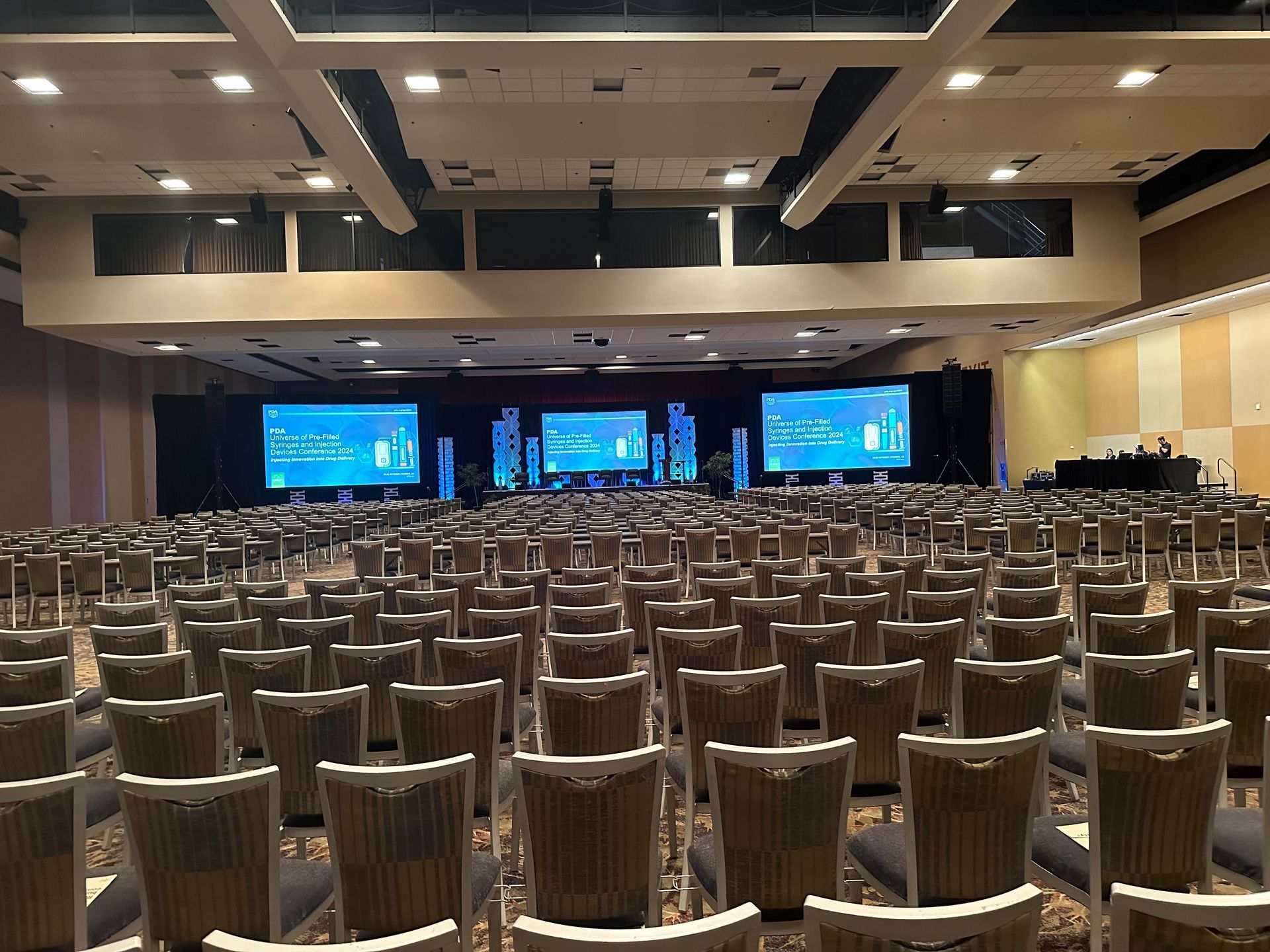 Conference hall with rows of empty chairs facing a stage with large blue presentation screens.