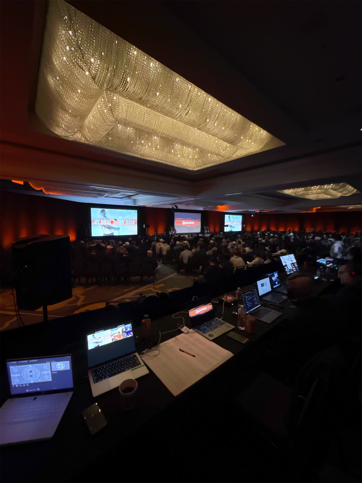 Large conference hall with audience seated at tables, multiple screens, and a bright patterned ceiling.